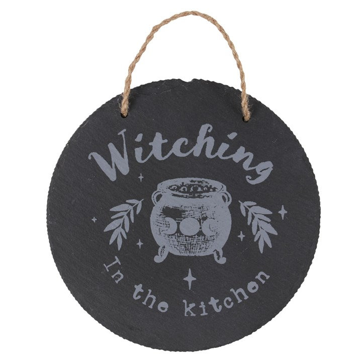 A circular slate sign with the words 'Witching in the Kitchen' on a white surface 