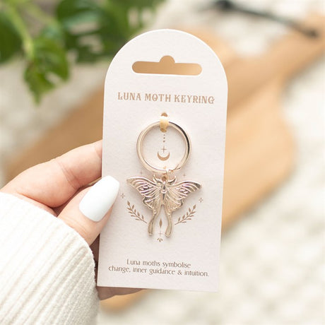 A light luna moth keyring with a dangling pendant, held on a card packaging with the text 'LUNA MOTH KEYRING' and a description of the product's meaning.