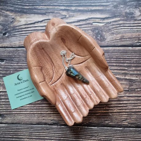 A Labradorite crystal pendulum displayed on a pair of wooden hands on a wooden surface next to an Arte's Den business card