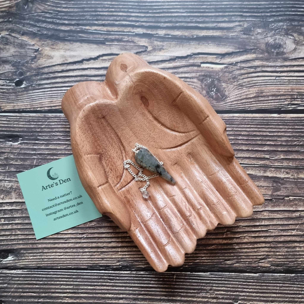 A Labradorite crystal pendulum displayed on a pair of wooden hands on a wooden surface next to an Arte's Den business card