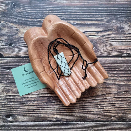 A flat point pendant with am Amazonite gemstone, laced into a black necklace with an adjustable string, displayed on a pair of wooden hands on a wooden surface next to a green Arte's Den business card 