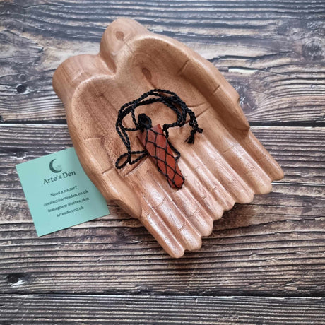 A flat point pendant with a red jasper gemstone, laced into a black necklace with an adjustable string, displayed on a pair of wooden hands on a wooden surface next to a green Arte's Den business card 