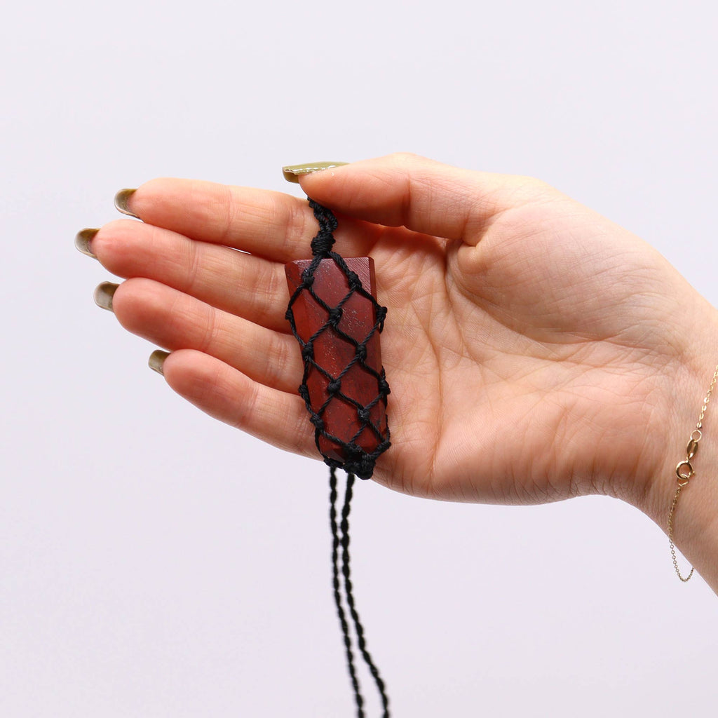 A flat point pendant with a red jasper gemstone, laced into a black necklace with an adjustable string, displayed in a hand