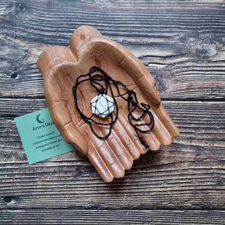 A hexagon pendant with a White Howlite gemstone, laced into a black necklace with an adjustable string, displayed on a pair of wooden hands on a wooden surface next to a green Arte's Den business card 