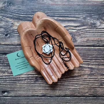 A hexagon pendant with a White Howlite gemstone, laced into a black necklace with an adjustable string, displayed on a pair of wooden hands on a wooden surface next to a green Arte's Den business card 