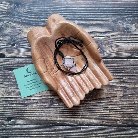 A teardrop shaped pendant with a Rose Quartz gemstone, laced into a black necklace with an adjustable string, displayed on a pair of wooden hands on a wooden surface next to a green Arte's Den business card 
