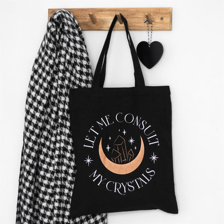 A black tote bag with crescent moon and crystal design which reads "Let me consult my crystals" hung up on a wooden coat hanger. 