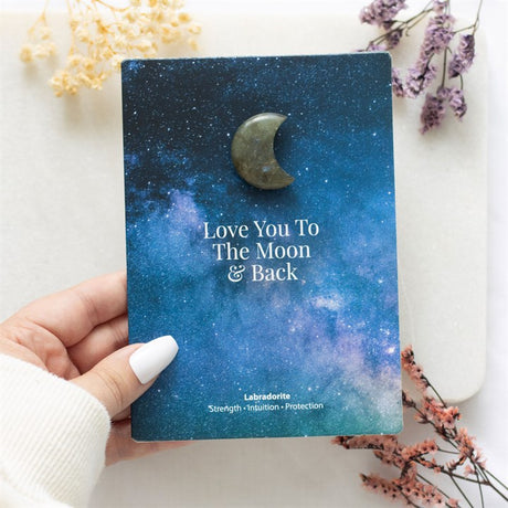 A hand holding a blue night sky themed card which says "Love you to the moon & back" with a Labradorite crescent moon stuck above it.