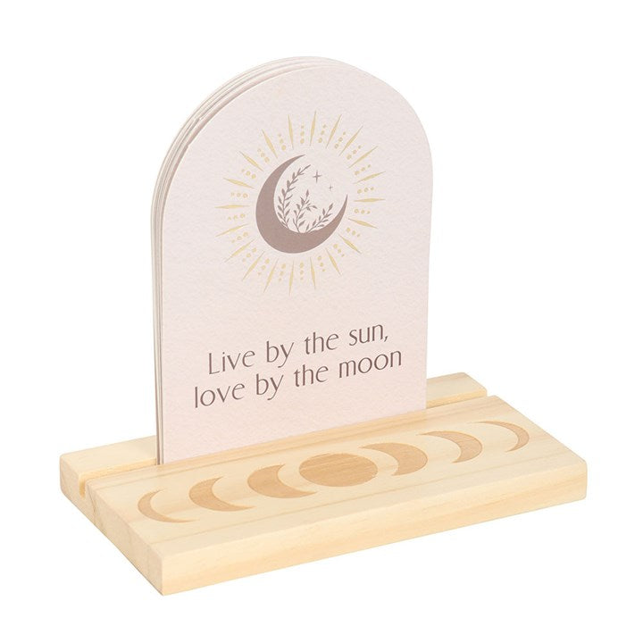 A beige affirmation card with moon phase design and the text 'Live by the sun, love by the moon' on it, placed on a wooden stand.