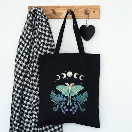 A black cotton tote bag with a mystical Luna Moth design, hanging on a wooden hook next to a houndstooth patterned jacket and decorative black heart
