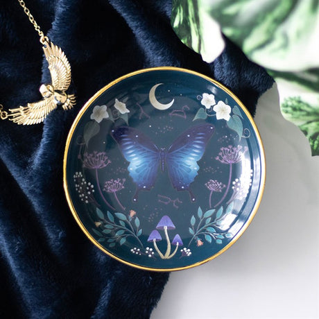 A round ceramic trinket dish with a moth design, gold edging, and floral elements, displayed against a blue background with a gold necklace.