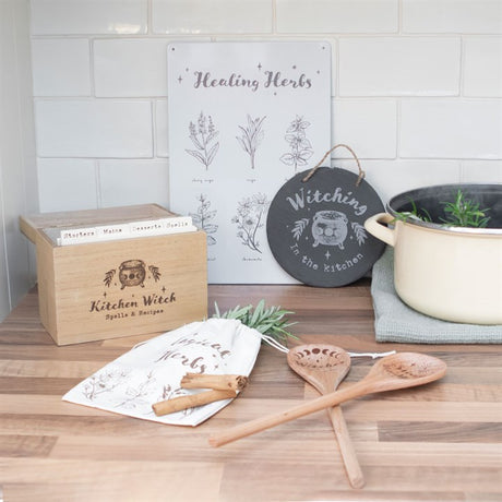 A circular slate sign with the words 'Witching in the Kitchen' hanging on a white wall above a wooden countertop, surrounded by kitchen-related items.