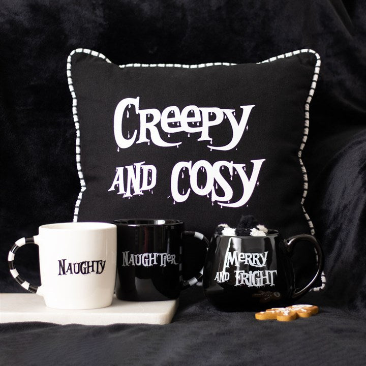 A black ceramic mug with 'Merry and Fright' text, placed on a black textured surface, with a pair of black and white striped socks inside it, next to a pair of "naughty and naughtier" mugs and a "creepy and cosy" cushion.