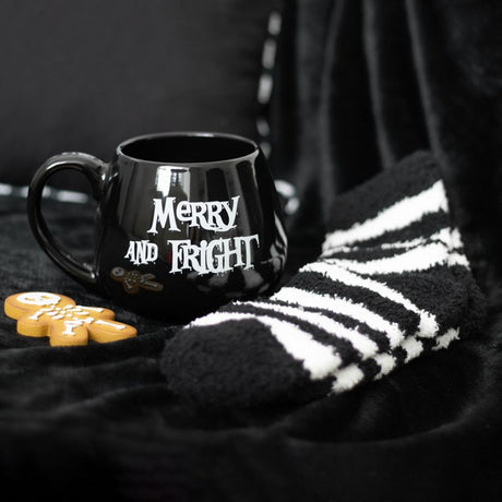 A black ceramic mug with 'Merry and Fright' text, placed on a black textured surface, with a pair of black and white striped socks beside it.