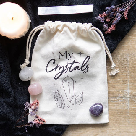 A beige cotton drawstring bag with 'My Crystals' text and a design of crystals printed on it, displayed on a dark fabric surface alongside some crystals and dried flowers.