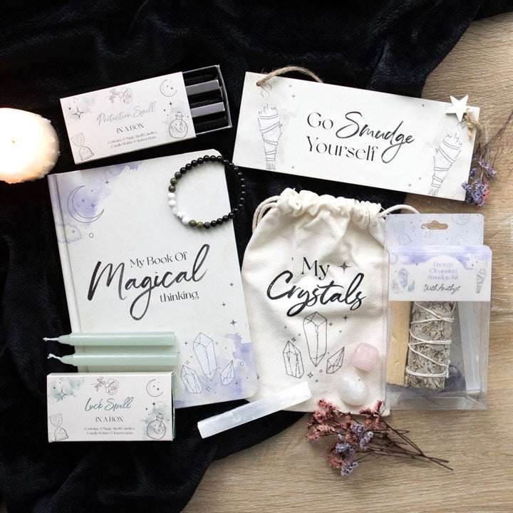 A selection of items including a beige cotton drawstring bag with 'My Crystals' text and a design of crystals printed on it, displayed on a dark fabric surface alongside some crystals, dried flowers and other items such as a notebook, bracelet and sign