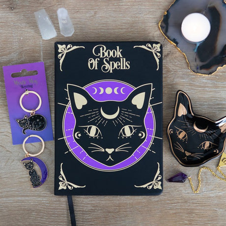 A black trinket dish with gold foil accents, shaped like a stylised cat face, displayed alongside a black book of spells and other trinkets.