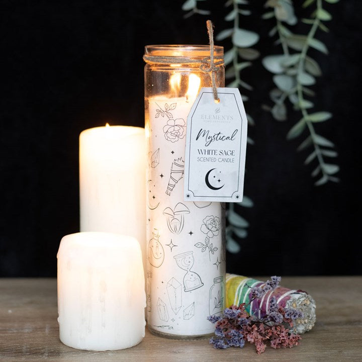 A "Mystical White Sage" glass tube candle with sage, flowers, and other decals, stands centre of the image next to lit candles on a wooden surface.