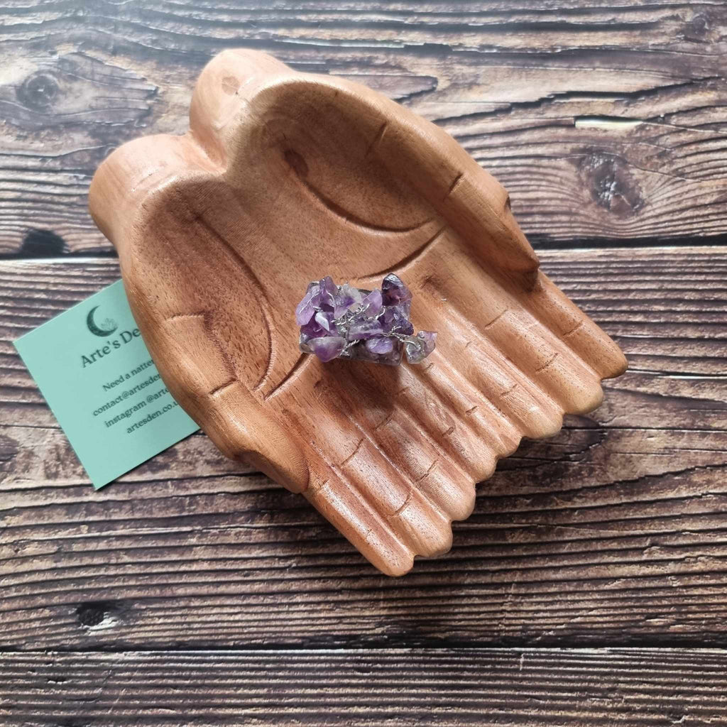 A mini amethyst tree on an orgonite base, displayed on a pair of wooden hands on a wooden surface with an Arte's Den business card