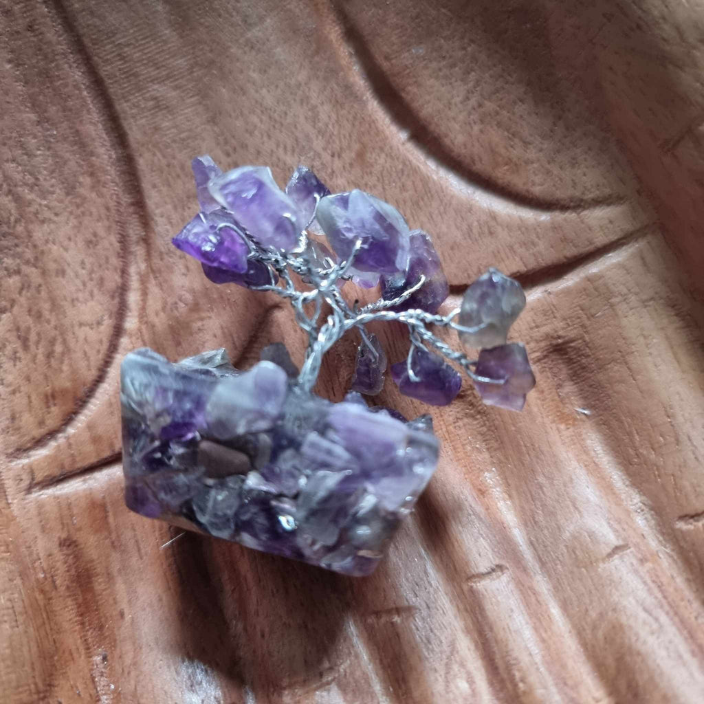 A mini amethyst tree on an orgonite base, displayed on a pair of wooden hands on a wooden surface with an Arte's Den business card