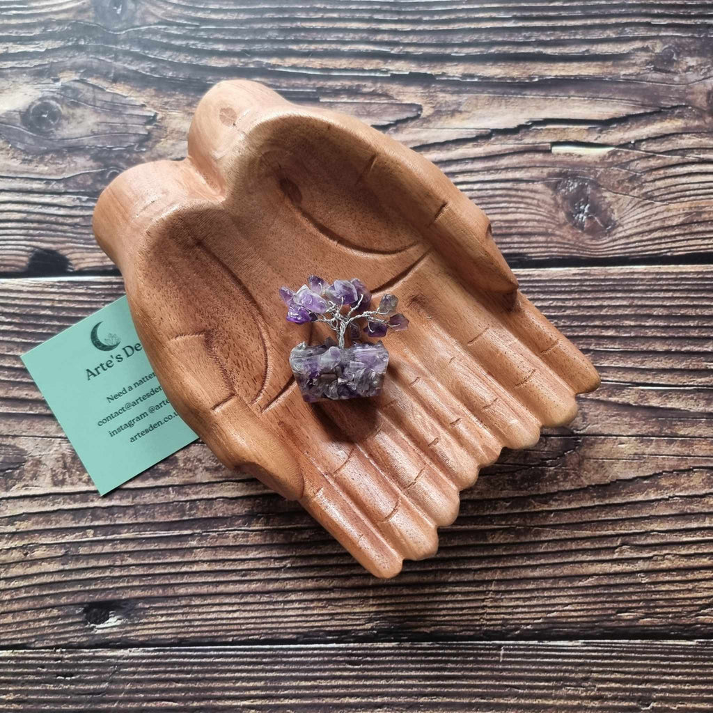 A mini amethyst tree on an orgonite base, displayed on a pair of wooden hands on a wooden surface with an Arte's Den business card