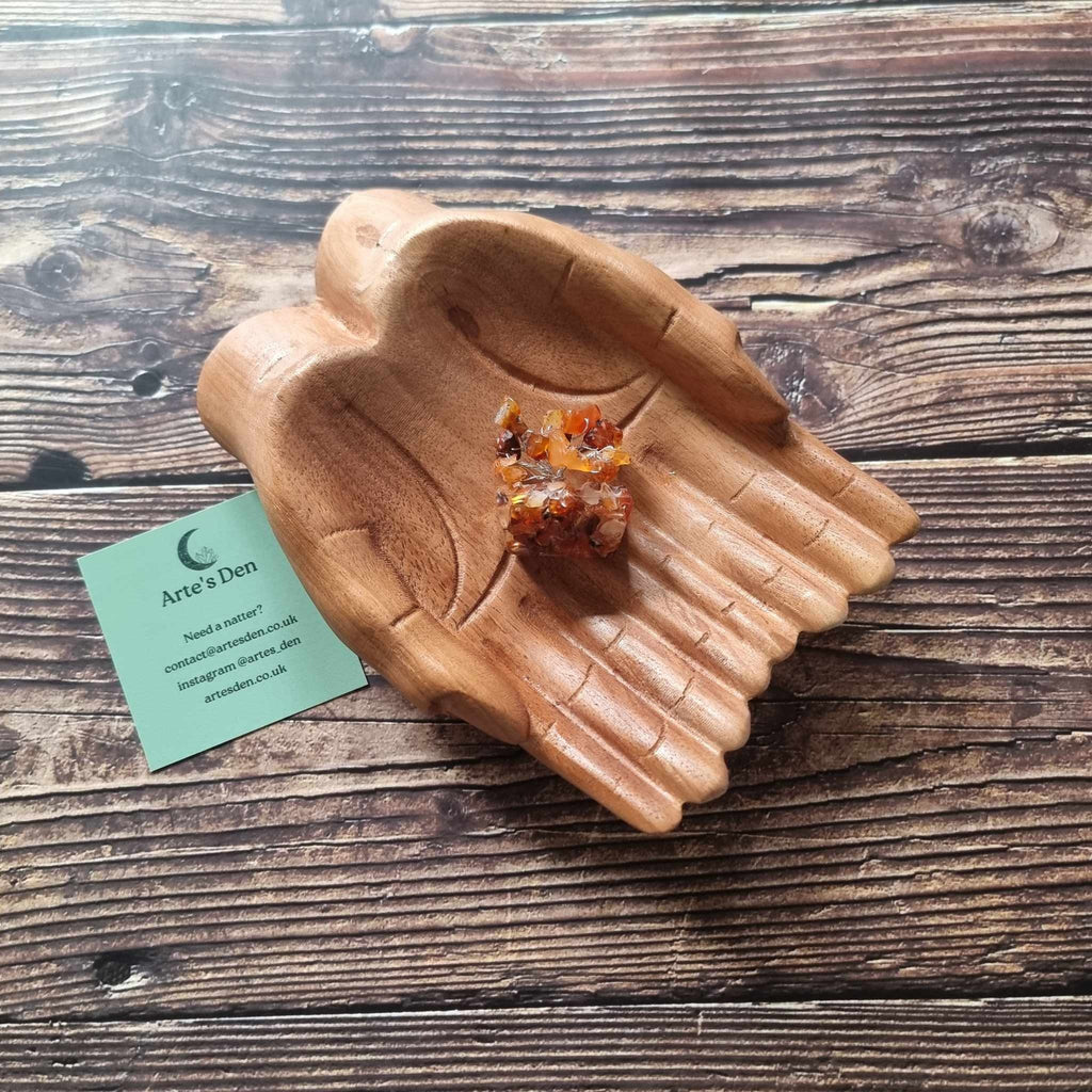 A mini carnelian tree on an orgonite base, displayed on a pair of wooden hands on a wooden surface with an Arte's Den business card
