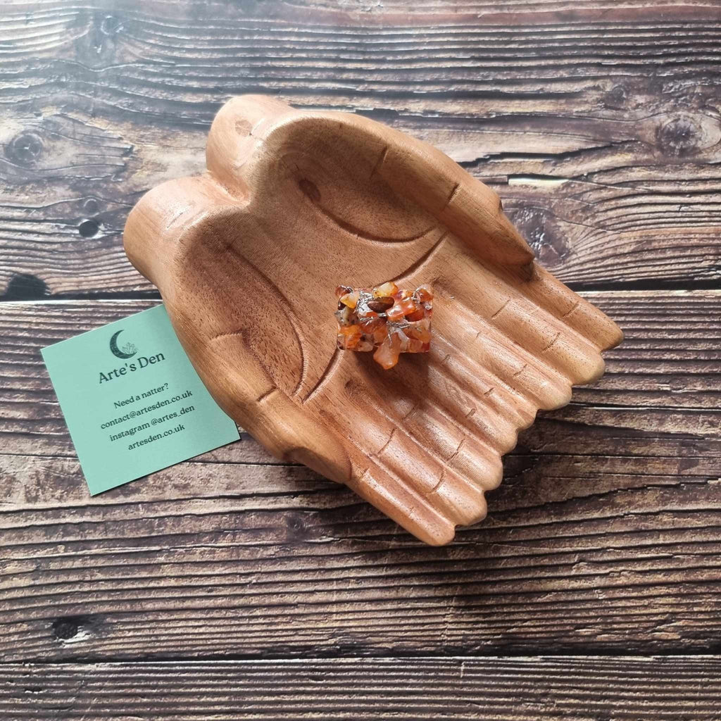 A mini carnelian tree on an orgonite base, displayed on a pair of wooden hands on a wooden surface with an Arte's Den business card
