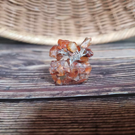 A mini carnelian tree on an orgonite base, with a translucent deep orangey-red appearance, placed against a woven background.