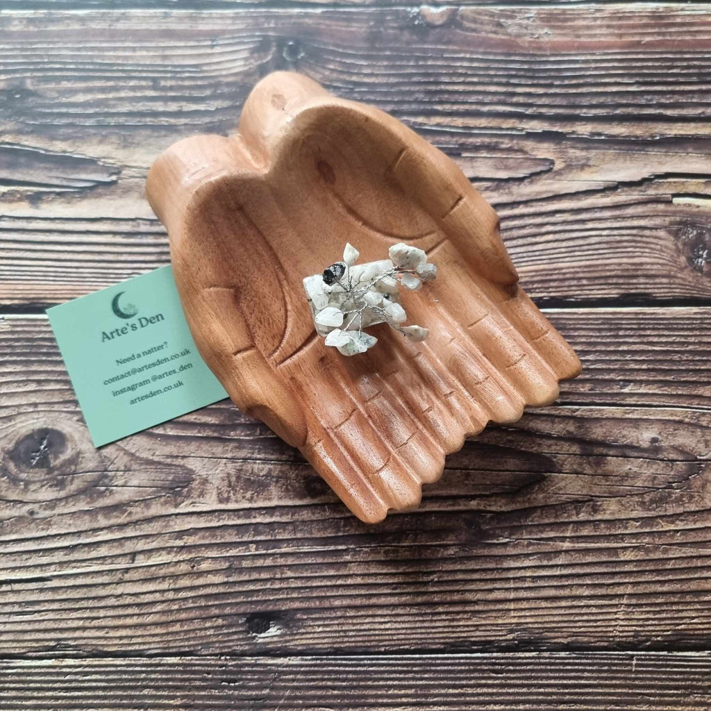 A mini white howlite tree on an orgonite base, displayed on a pair of wooden hands on a wooden surface with an Arte's Den business card