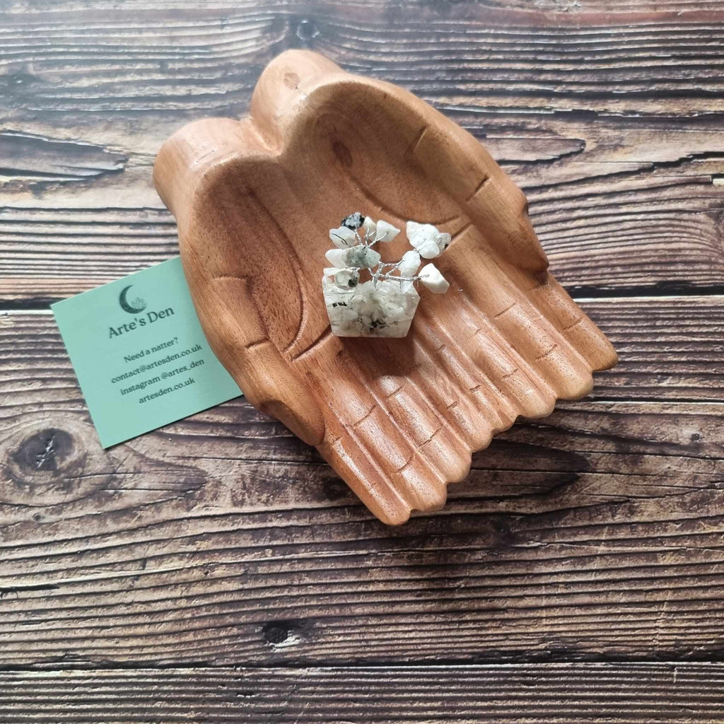 A mini white howlite tree on an orgonite base, displayed on a pair of wooden hands on a wooden surface with an Arte's Den business card