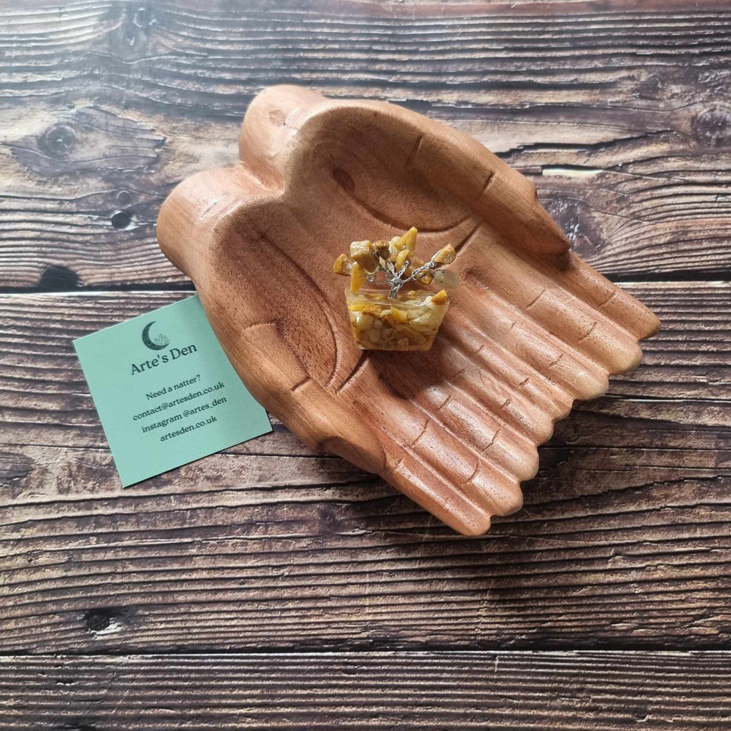 A mini yellow jasper tree on an orgonite base, displayed on a pair of wooden hands on a wooden surface with an Arte's Den business card