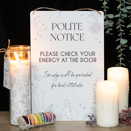 A tin sign with an illustration of crystals, potion bottles, and smudge sticks, accompanied by the phrase 'POLITE NOTICE PLEASE CHECK YOUR ENERGY AT THE DOOR. SMOUDGE WILL BE PROVIDED FOR BAD ATTITUDES. It is beside lit candles.