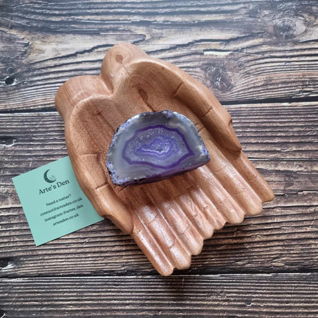 A purple agate geode displayed in a wooden hand-shaped dish with a business card beside it, on a wooden background.