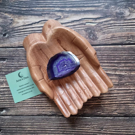 A purple agate geode displayed in a wooden hand-shaped dish with a business card beside it, on a wooden background.