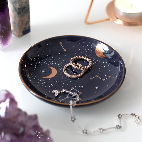 A ceramic trinket dish with a purple background featuring celestial patterns, including stars and constellations, displayed on a table with jewellery on top.