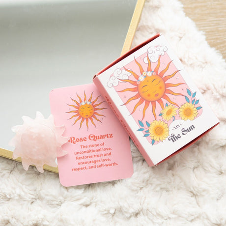 A carved Rose Quartz crystal sun alongside its packaging, which includes a card with the text 'Rose Quartz' and 'The Sun' indicating a tarot card-inspired design. The items are presented on a soft, fluffy surface.