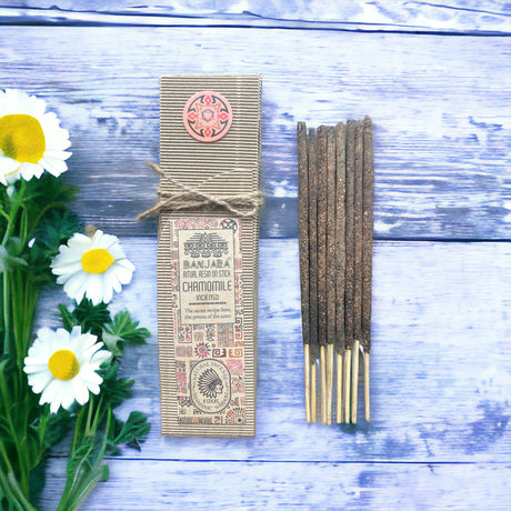 Ritual Incense sticks laid on a wooden backdrop next to white and yellow flowers. Also next to its beige packaging tied with string. 