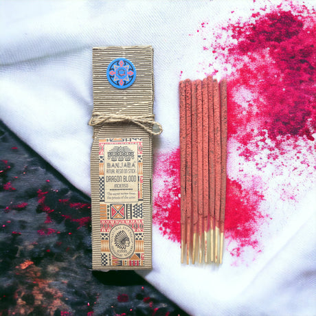 Ritual Incense sticks laid on a white background with splashes of pink next to its beige packaging which is tied with string. 