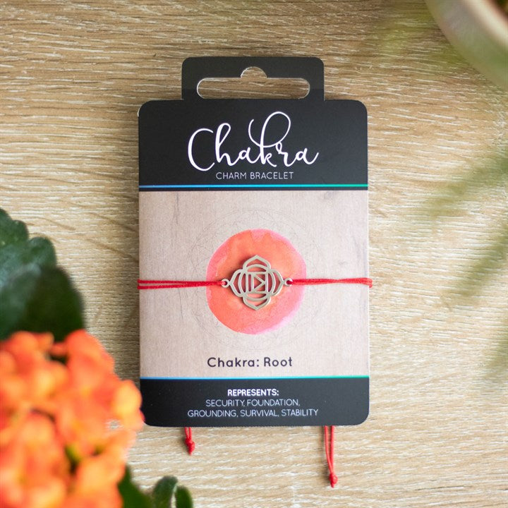 A Root Chakra charm bracelet with a red band and a silver charm, displayed on a card with 'Chakra' written on it, placed on a wooden surface with orange flowers in the background.