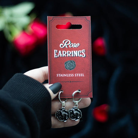 A pair of silver stainless steel rose earrings dangling from a card with a red background, held by a hand.