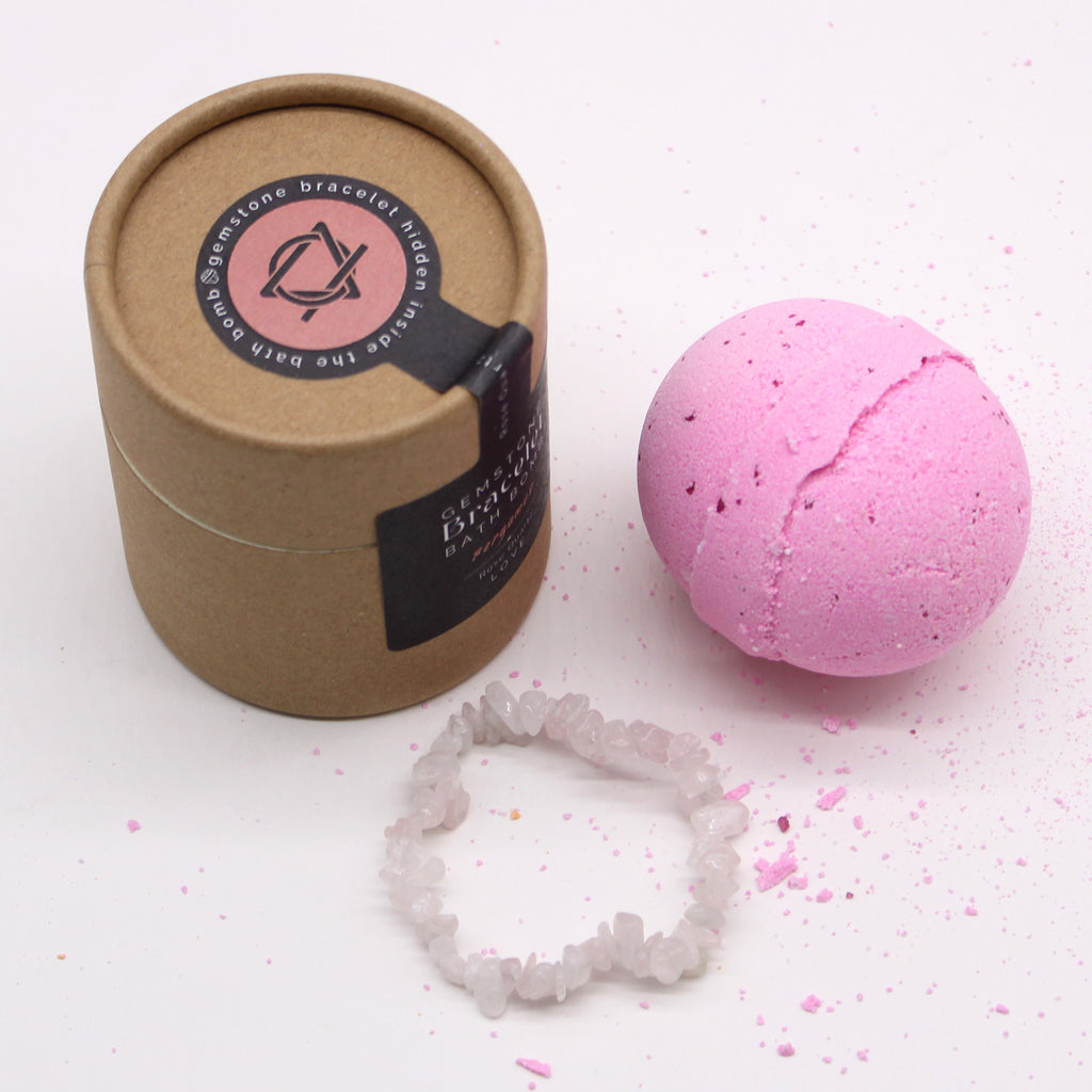 A brown package with black label stating "gemstone bracelet bath bomb" next to a pink bath bomb and rose quartz bracelet on a white background. 