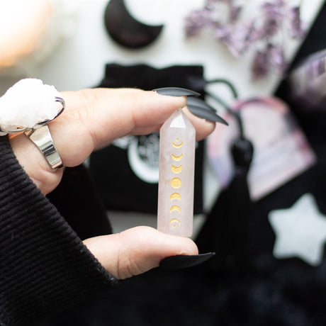 A hand holding a Rose Quartz crystal point with gold-tone moon phase engraving, with a blurred background.