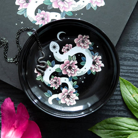 A round, black trinket dish with a white snake and pink flower design, placed on a dark surface with a starry background. The dish has a chain laid on it, and there are decorative leaves and a pink flower in the foreground.