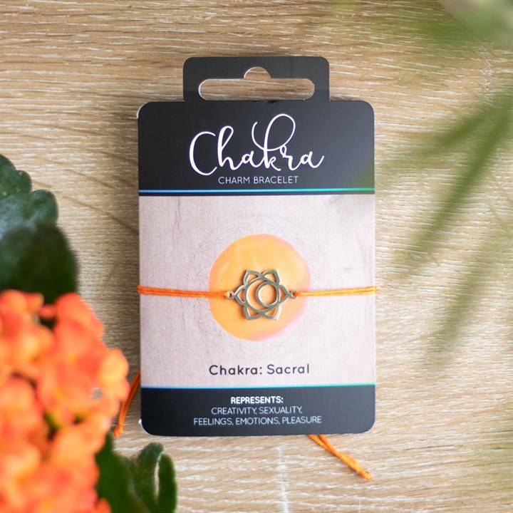 A Sacral Chakra charm bracelet with an orange band and a silver charm, displayed on a card with 'Chakra' written on it, placed on a wooden surface with orange flowers in the background.