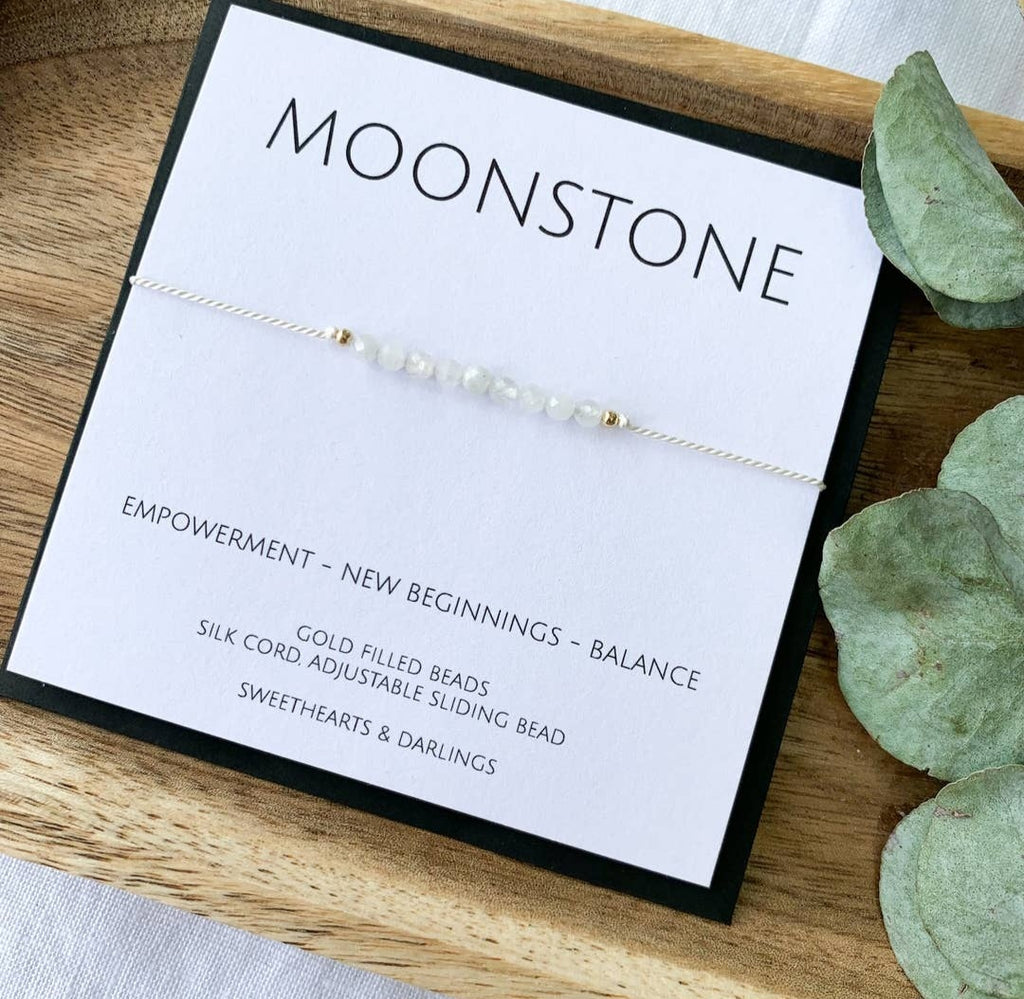 A Moonstone silk bracelet with beads approximately 3mm in size, displayed on a wooden tray alongside a keepsake card that reads 'Moonstone - EMPOWERMENT - NEW BEGINNINGS - BALANCE'.