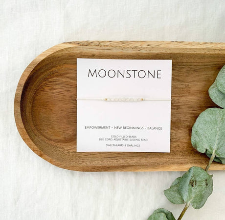 A Moonstone silk bracelet with beads approximately 3mm in size, displayed on a wooden tray alongside a keepsake card that reads 'Moonstone - EMPOWERMENT - NEW BEGINNINGS - BALANCE'.