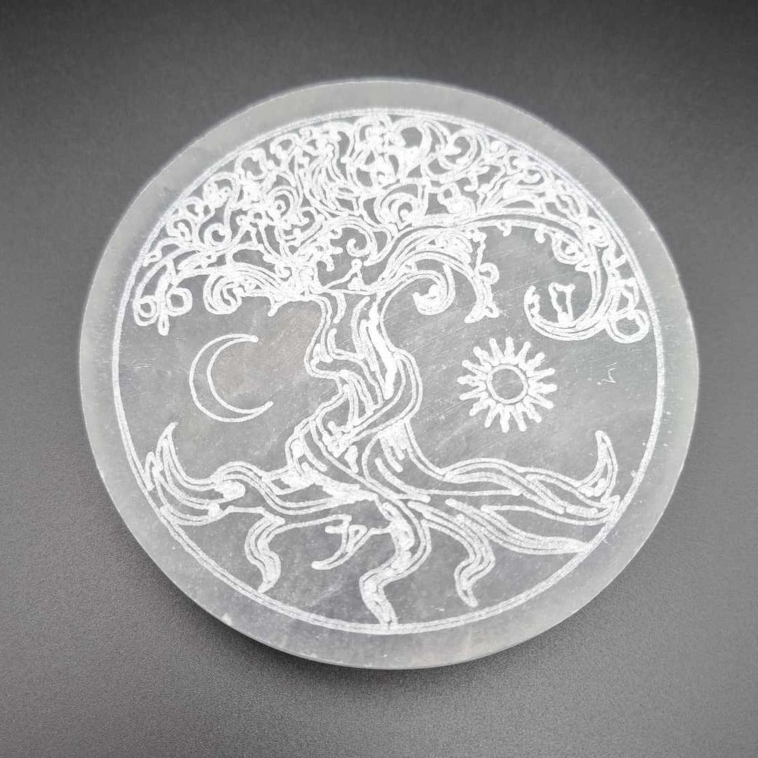 A Selenite charging plate which is round, milky white and has an etched Tree of Life design along with a crescent moon and sun.