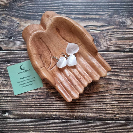 Three Selenite heart pendants sit inside a pair of wooden hands on a wooden surface next to a green Arte's Den business card 