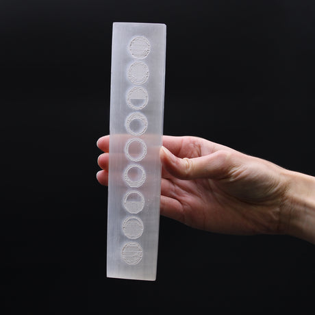Hand holding a selenite charging bar with moon phases design against a black background