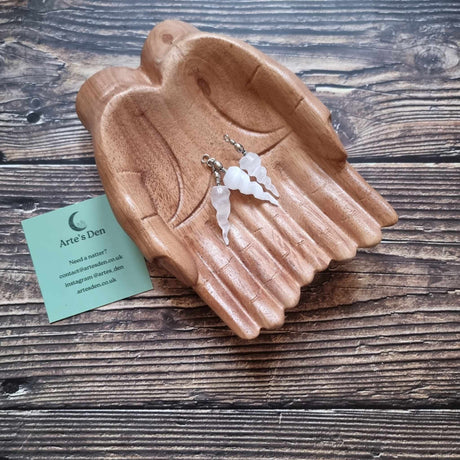 Three Selenite spiral pendants sit inside a pair of wooden hands on a wooden surface next to a green Arte's Den business card 